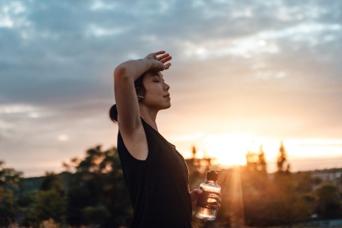 A woman stands outdoors at sunset, shielding her eyes with one hand and holding a water bottle in the other.