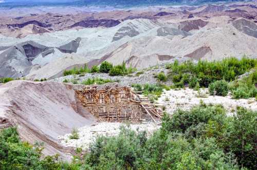 A landscape of colorful, layered hills with a wooden structure partially hidden among greenery in the foreground.