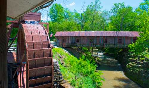 A wooden water wheel beside a creek, with a rustic covered bridge and lush greenery in the background.