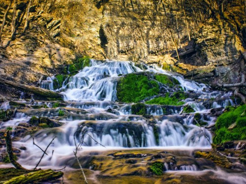 A serene waterfall cascades over rocky terrain, surrounded by lush greenery and sunlit cliffs.