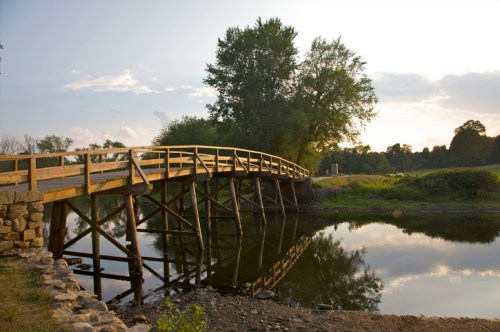 A wooden bridge spans a calm river, surrounded by trees and a serene landscape under a cloudy sky.
