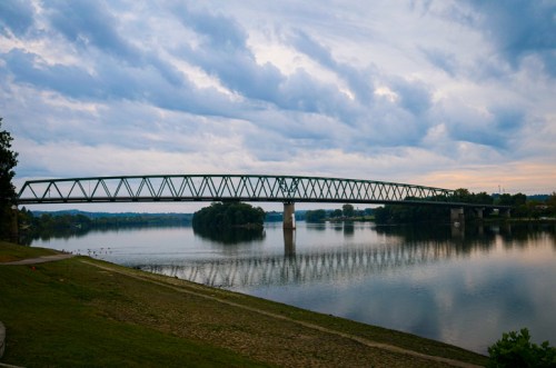 A green bridge spans a calm river, reflecting the cloudy sky and surrounding trees at dusk.