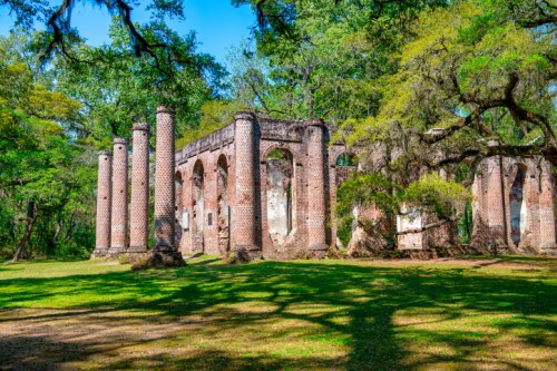 Ruins of a historic brick structure surrounded by lush greenery and trees under a clear blue sky.