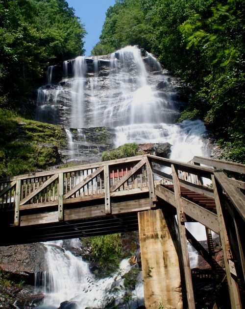 A wooden bridge in front of a cascading waterfall surrounded by lush green trees under a clear blue sky.