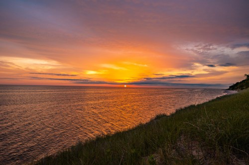 A vibrant sunset over a calm ocean, with grassy shoreline and colorful clouds reflecting on the water.