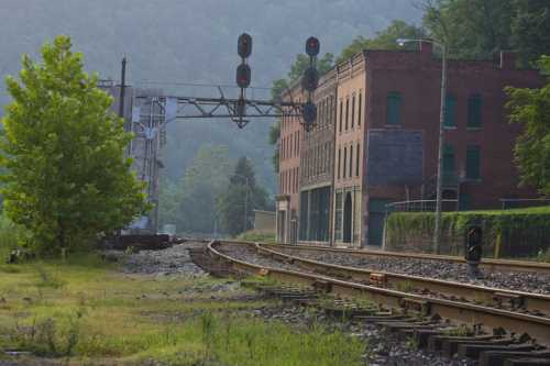 A quiet railway scene featuring tracks, a signal bridge, and an old brick building surrounded by greenery.