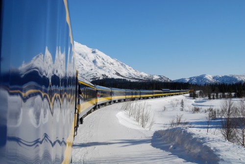 A train winds through a snowy landscape with mountains under a clear blue sky.