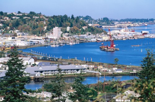 A scenic view of a coastal town with a harbor, boats, and surrounding greenery under a clear blue sky.