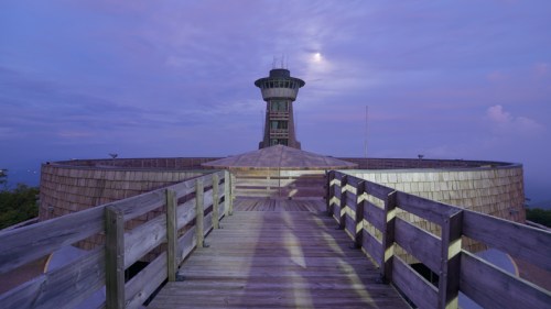 A wooden walkway leads to a circular tower at dusk, surrounded by a serene landscape under a purple sky.