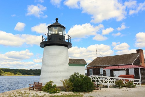 A white lighthouse beside a calm waterway, with a nearby building and a bench under a blue sky with fluffy clouds.