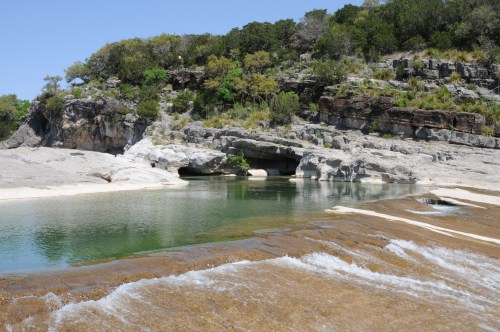 A serene river scene with clear water flowing over rocks, surrounded by lush greenery and rugged cliffs.