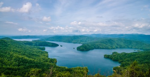 A panoramic view of a lush green landscape surrounding a serene lake under a partly cloudy sky.