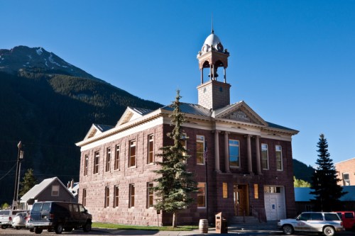 Historic stone building with a clock tower, surrounded by trees and mountains under a clear blue sky.