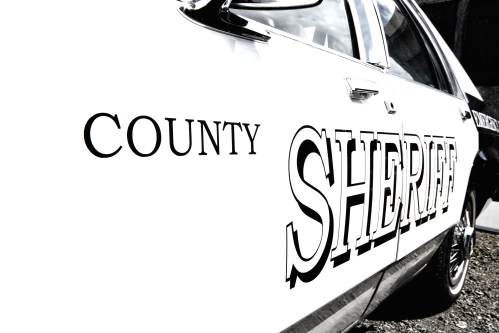 Close-up of a white sheriff's car with "COUNTY SHERIFF" printed on the side in bold black letters.