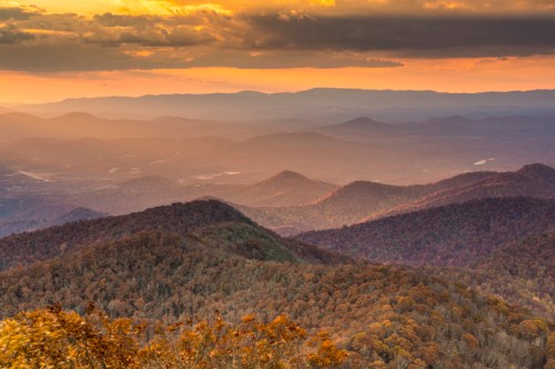 A scenic view of rolling mountains at sunset, with warm hues illuminating the landscape and autumn foliage.