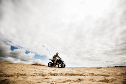 A person riding an ATV on sandy terrain, with a flag attached, under a cloudy sky.