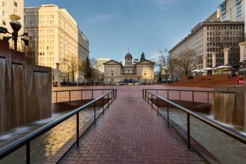 A plaza with brick pathways, water features, and buildings in the background under a clear blue sky.