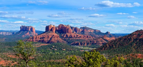 A scenic view of red rock formations and green trees under a blue sky with fluffy clouds in Sedona, Arizona.