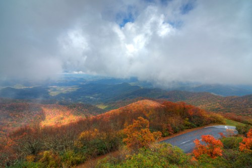 A scenic view of colorful autumn foliage on mountains under a cloudy sky, with a winding road in the foreground.