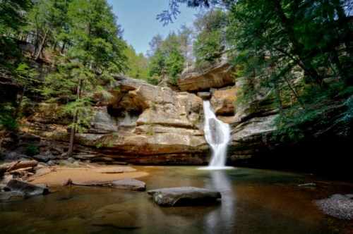 A serene waterfall cascades into a calm pool, surrounded by lush green trees and rocky cliffs.