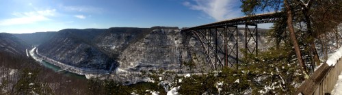 A panoramic view of a snowy landscape featuring a bridge over a river, surrounded by steep, snow-covered hills.