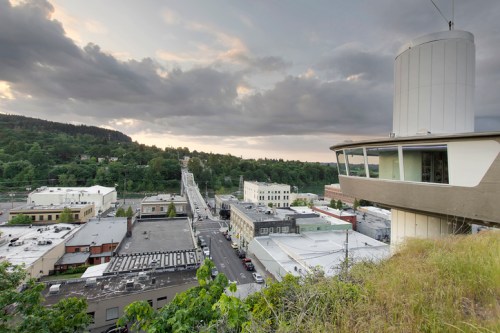 A panoramic view of a town with buildings, a bridge, and lush greenery under a cloudy sky.
