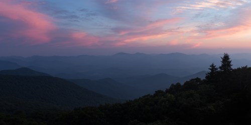A panoramic view of misty mountains at sunset, with soft pink and purple clouds in the sky.