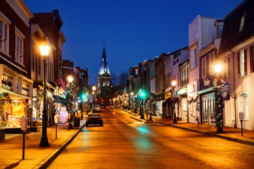 A quiet street at dusk, lined with shops and festive lights, leading to a church steeple in the distance.