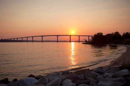 Sunset over a calm river, with a bridge in the background and rocky shoreline in the foreground.