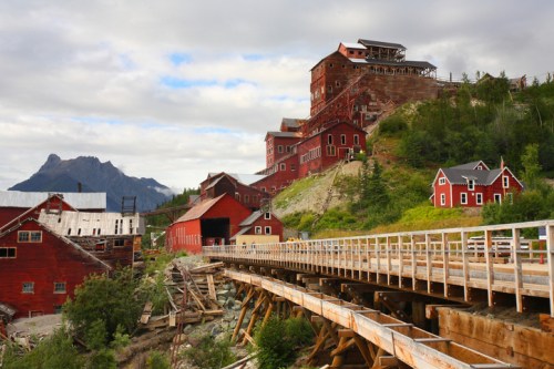 Historic red buildings on a hillside, with a wooden walkway and mountains in the background under a cloudy sky.