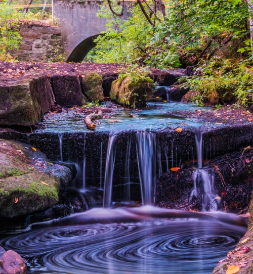 A serene waterfall cascades over rocks, surrounded by lush greenery and a peaceful stream under a stone bridge.