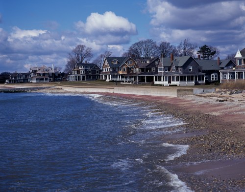 A shoreline with gentle waves lapping at the sandy beach, lined by charming houses under a cloudy sky.