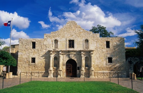 The Alamo, a historic stone mission, with a Texas flag and blue sky in the background.