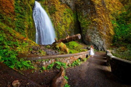 A woman in a white dress stands near a waterfall surrounded by lush greenery and moss-covered rocks.