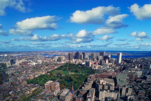 Aerial view of a city skyline with green parks, buildings, and a blue sky dotted with clouds.