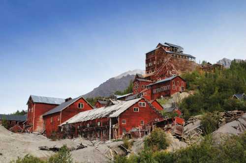 Abandoned red mining buildings nestled in a mountainous landscape under a clear blue sky.