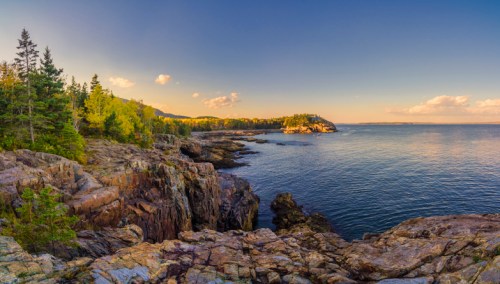 A scenic coastal view featuring rocky cliffs, lush trees, and calm waters under a clear blue sky.