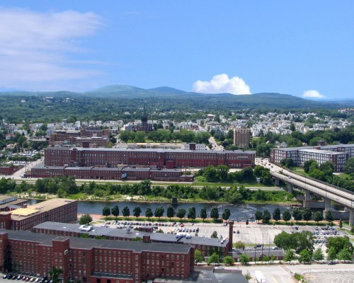 Aerial view of a city with industrial buildings, a river, and mountains in the background under a clear blue sky.
