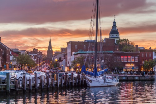 A scenic waterfront view at sunset, featuring a sailboat, historic buildings, and a church steeple in the background.