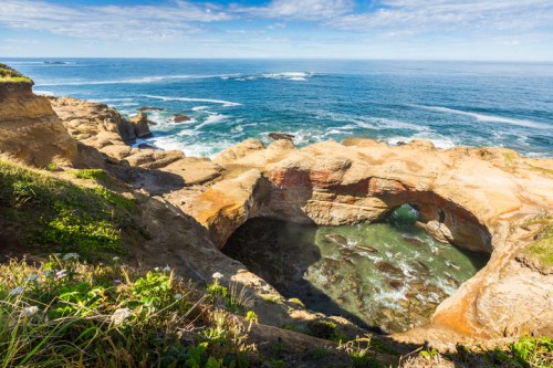 A scenic coastal view featuring a rocky cliff, a natural arch, and clear blue waters under a bright sky.
