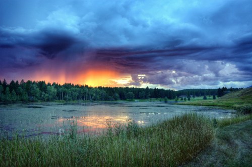 A serene lake at sunset, surrounded by lush greenery and dramatic clouds in vibrant colors.