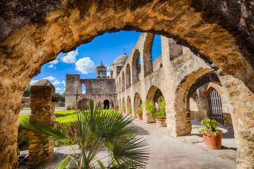 Historic stone arches frame a lush courtyard with greenery and a blue sky, showcasing a serene, ancient structure.