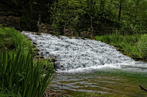 A serene waterfall cascades over stone steps, surrounded by lush greenery and a calm pool below.