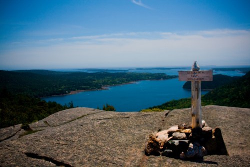 A scenic view from a mountain summit overlooking a lake and lush green landscape, with a wooden signpost in the foreground.