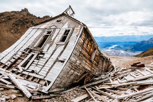 A tilted, abandoned wooden house on a rocky hillside, surrounded by scattered debris and a distant valley view.