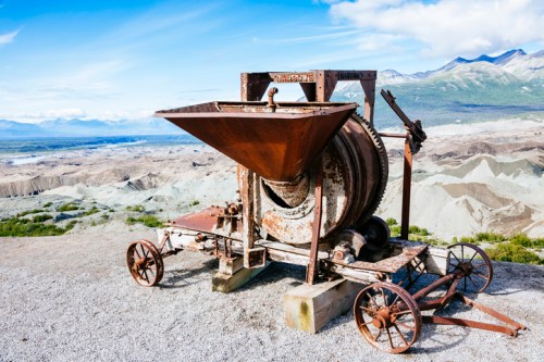 An old, rusted machine sits on a gravel surface with a mountainous landscape in the background under a blue sky.