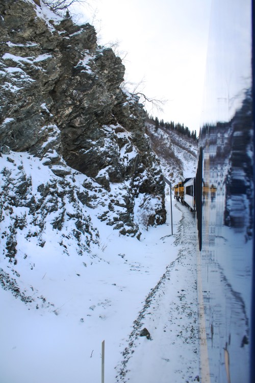 A train travels along a snowy path beside rocky cliffs in a winter landscape.