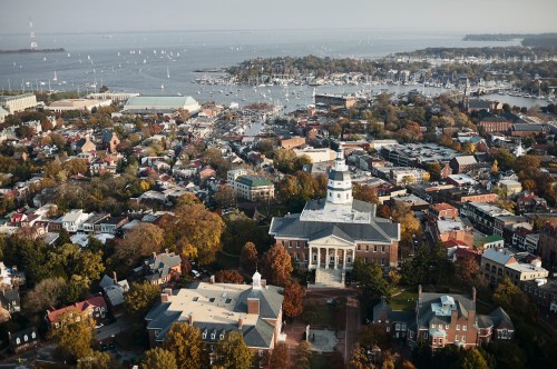 Aerial view of a coastal town with boats, waterfront, and historic buildings surrounded by autumn foliage.