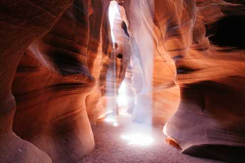 A narrow canyon with smooth, reddish walls and beams of light illuminating the sandy floor.