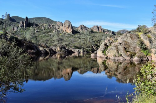 A serene landscape featuring a calm lake reflecting rocky hills and greenery under a clear blue sky.